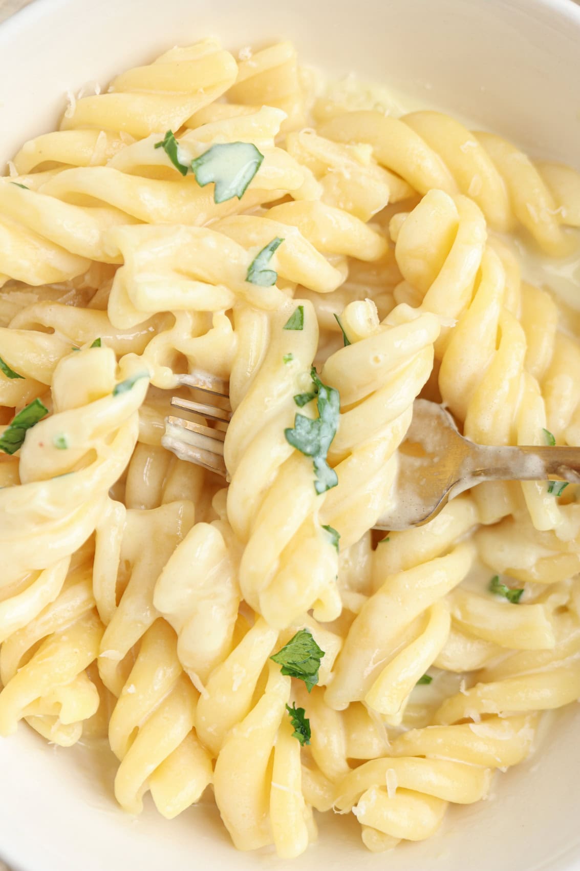 A close-up of creamy pasta spirals garnished with chopped fresh herbs, with a fork twirling some of the pasta in a white bowl.