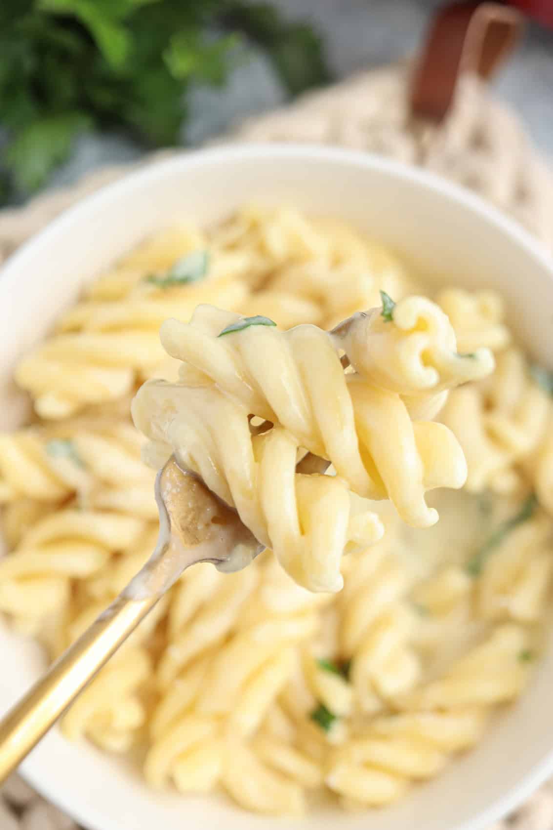 A close-up of a fork holding creamy cottage cheese alfredo pasta rotini above a bowl filled with more pasta, garnished with small pieces of parsley.