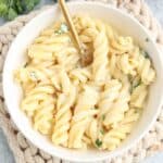 A white bowl filled with creamy alfredo pasta, garnished with chopped parsley, and a gold fork resting inside. The bowl is placed on a beige woven mat.