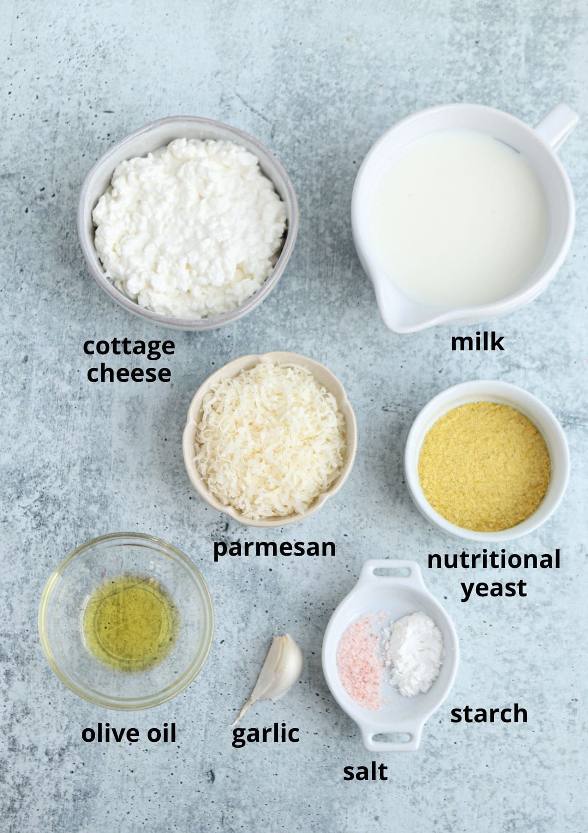 A flat lay of labeled ingredients for ricotta pesto pasta on a gray surface: cottage cheese, milk, parmesan, nutritional yeast, olive oil, garlic clove, a small dish of starch, and a small dish with pink salt.
