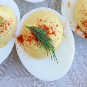 A close-up of a deviled egg topped with paprika and a small sprig of dill, placed on a light-colored patterned surface.