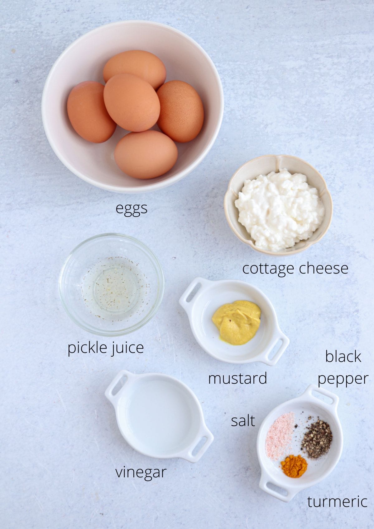 A flat lay of ingredients in small bowls, labeled as: eggs, cottage cheese, pickle juice, mustard, black pepper, vinegar, salt, and turmeric—arranged on a light-colored surface.