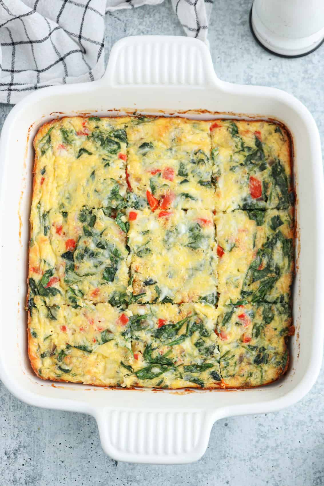 A cottage cheese egg bake with spinach and red bell peppers is cut into six squares, sitting in a white square baking dish on a light gray surface. A checkered kitchen towel is partially visible in the top left corner.