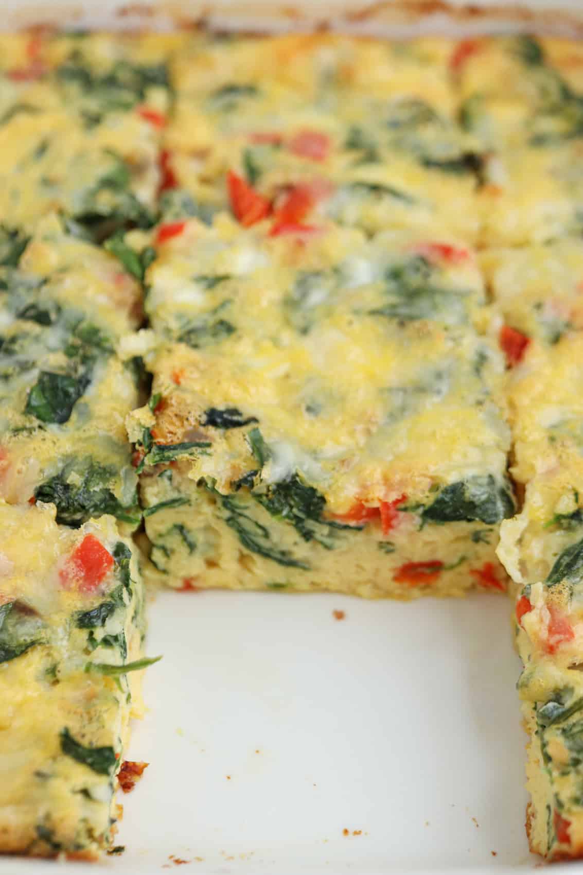 A close-up of a cottage cheese egg bake casserole with visible pieces of spinach, red bell pepper, and melted cheese, cut into square pieces in a white baking dish. One square has been removed from the dish.