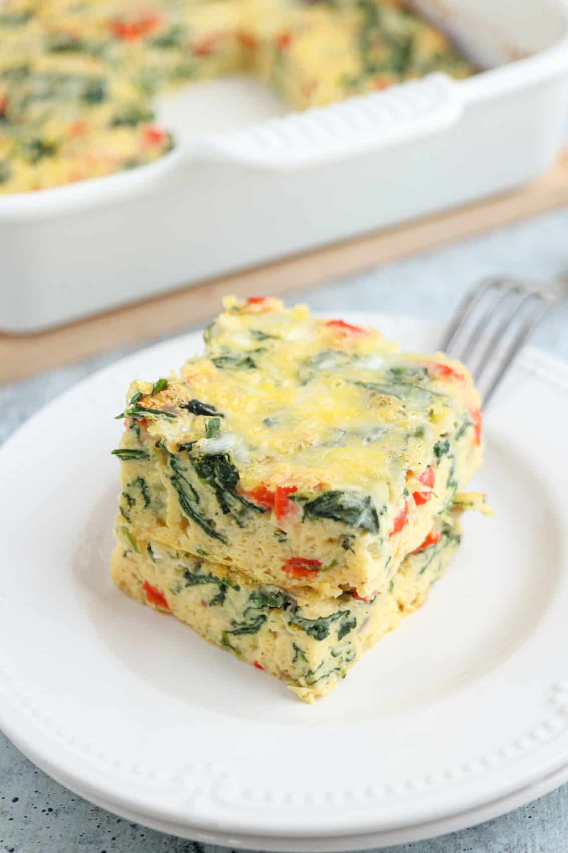 Two square pieces of cottage cheese egg bake with spinach and red peppers are stacked on a white plate, with a fork beside them and a baking dish of casserole in the background.