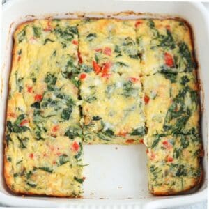 A cottage cheese egg bake featuring chopped spinach and red bell peppers, sliced into squares in a white baking dish, with one piece missing from the center front.