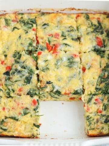 A cottage cheese egg bake featuring chopped spinach and red bell peppers, sliced into squares in a white baking dish, with one piece missing from the center front.