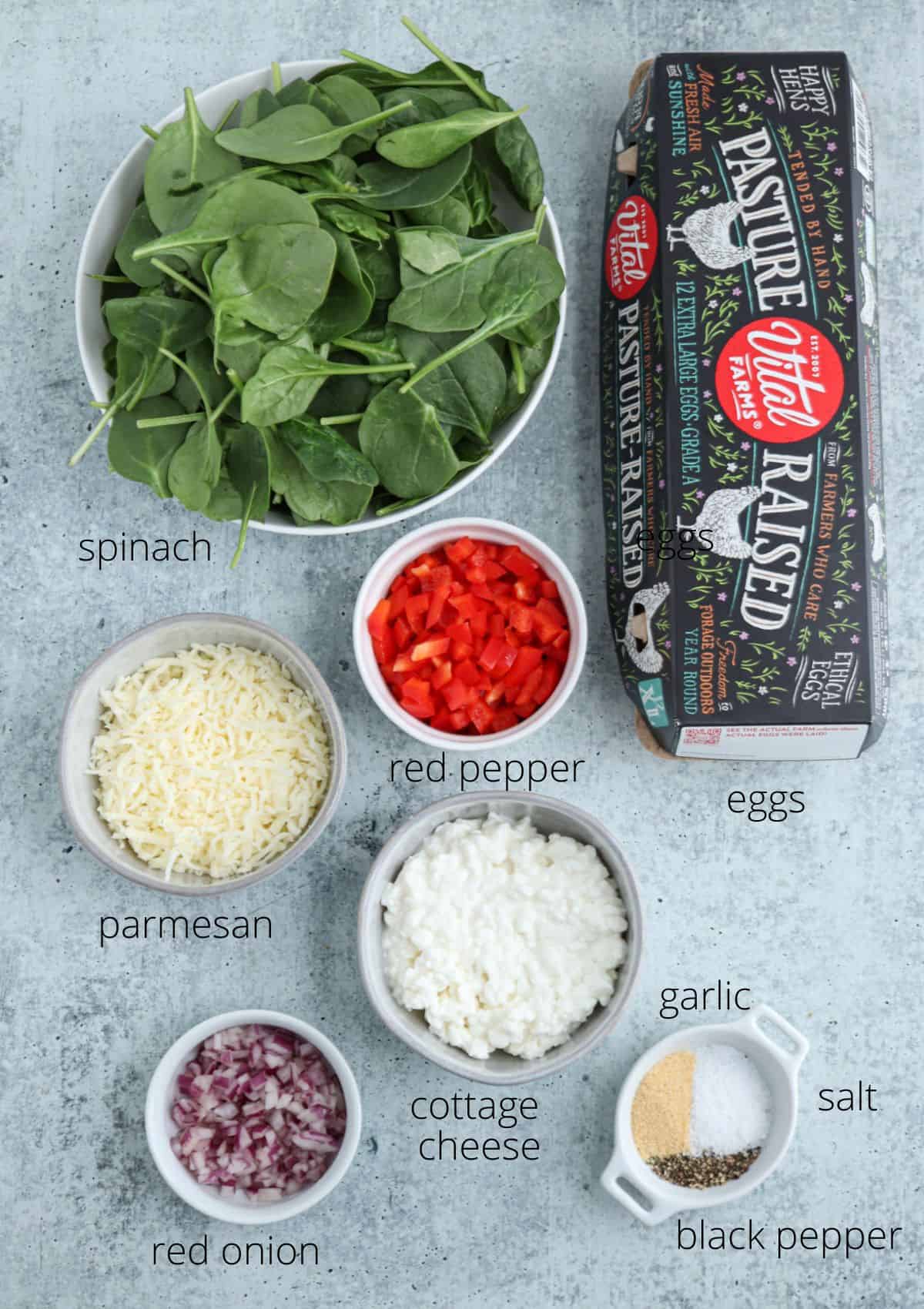 A carton of eggs, a bowl of fresh spinach, chopped red pepper, shredded parmesan, cottage cheese, and other ingredients for a tasty cottage cheese egg bake are arranged on a light surface with ingredient labels.