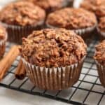 A close-up of a spiced muffin with an oat topping, sitting on a cooling rack with other similar muffins in the background. Two cinnamon sticks are placed beside the muffin.
