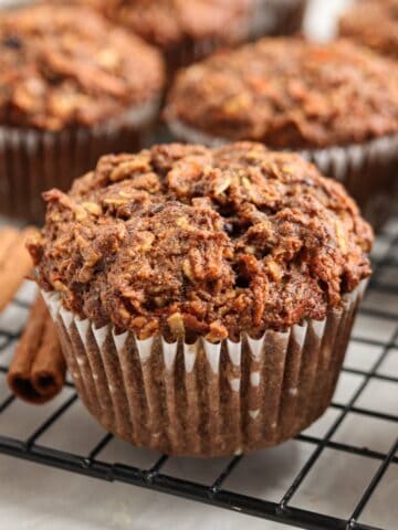A close-up of a spiced muffin with an oat topping, sitting on a cooling rack with other similar muffins in the background. Two cinnamon sticks are placed beside the muffin.