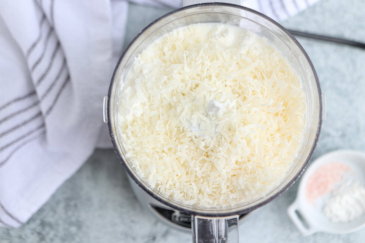 A food processor bowl filled with shredded cheese and creamy ingredients for cottage cheese alfredo sits on a light surface beside a striped white towel and a small dish containing pink salt and cornstarch.