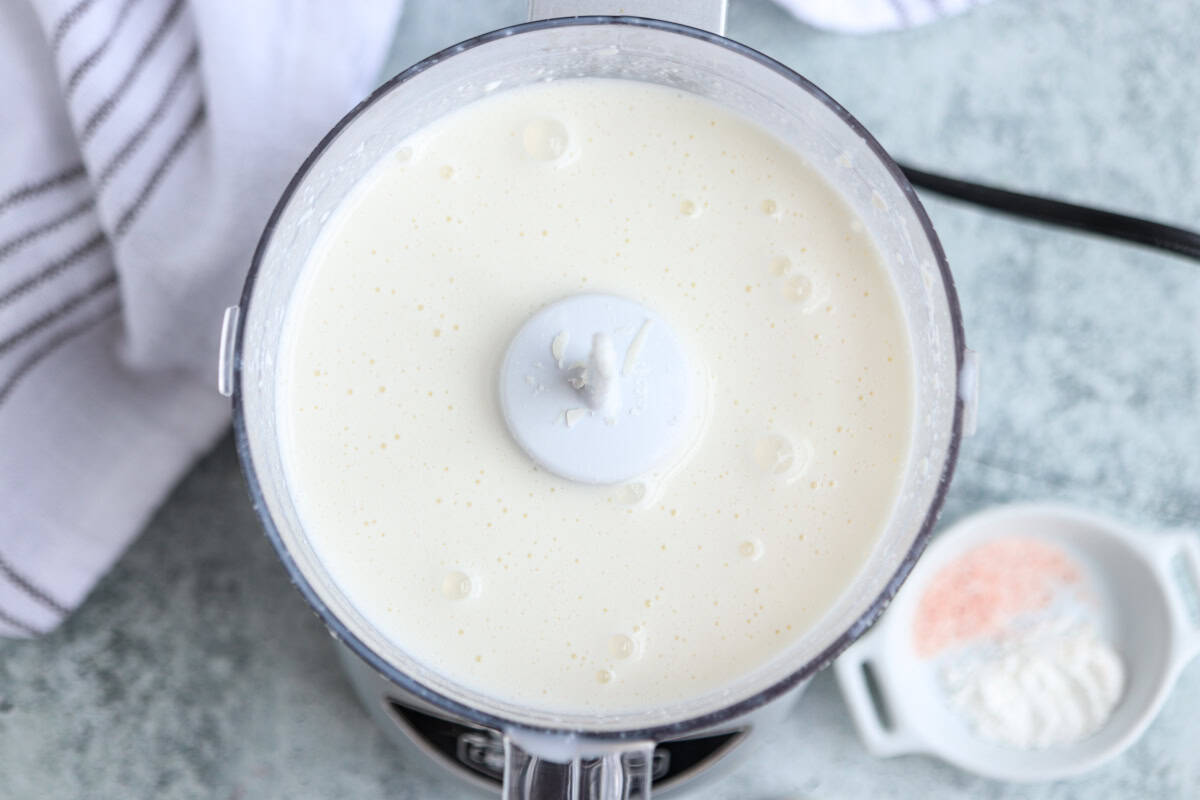 Overhead view of a blender filled with a smooth, creamy white mixture. A white towel with grey stripes and a small dish holding pink salt and flour are nearby on a light-colored surface.