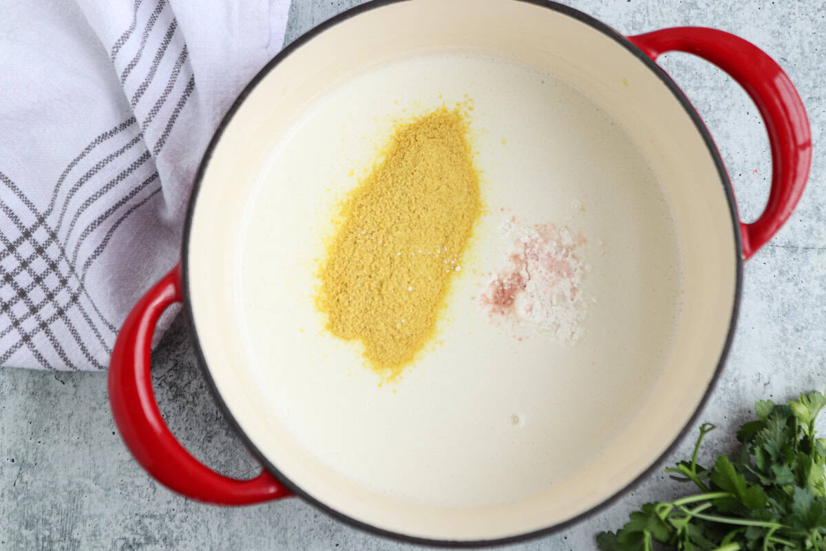 A white pot with red handles contains a creamy cottage cheese alfredo, topped with yellow and pink powders. A white towel with black stripes and some fresh cilantro are nearby on a light countertop.