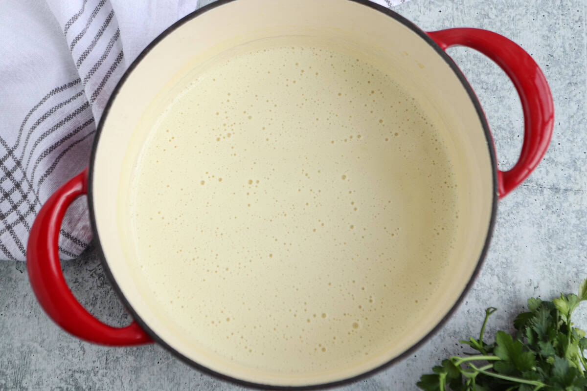 Overhead view of a red-handled pot filled with creamy sauce, sitting on a gray surface. A white kitchen towel and fresh herbs are partially visible beside the pot.