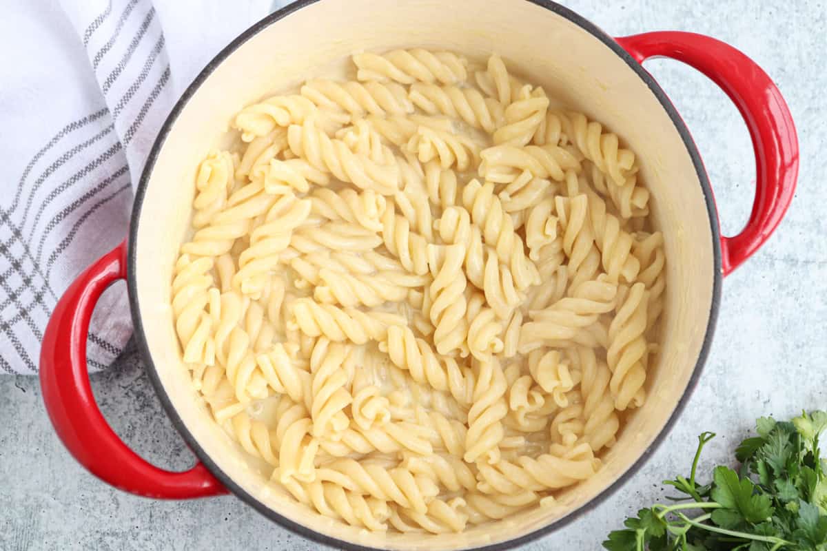 A red-handled pot filled with creamy cottage cheese alfredo fusili, sits on a light surface next to a white towel with gray stripes and a sprig of fresh parsley.