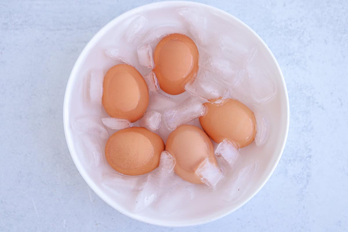 Five brown hard-boiled eggs cooling in an ice bath inside a white bowl, placed on a light gray surface.