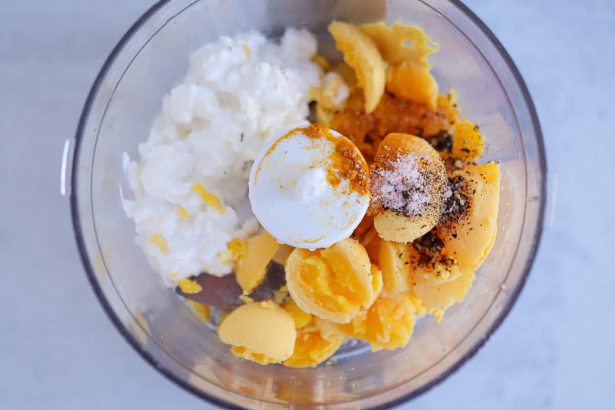 Overhead view of a food processor bowl with cottage cheese, cooked egg yolks, cottage cheese, and spices like turmeric.
