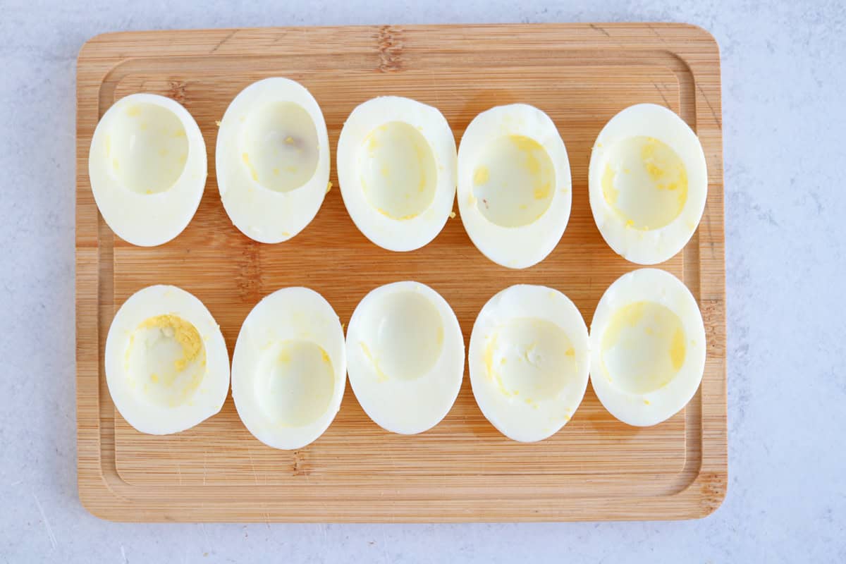 Ten hard-boiled egg halves with yolks removed are neatly arranged in two rows on a rectangular wooden cutting board on a light-colored surface.