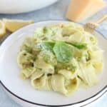 A plate of wide ricotta pesto pasta noodles topped with green pesto sauce and fresh basil leaves, served on a white plate with a gold fork and a wedge of cheese in the background.