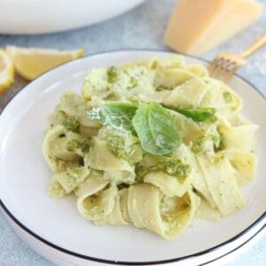 A plate of wide ricotta pesto pasta noodles topped with green pesto sauce and fresh basil leaves, served on a white plate with a gold fork and a wedge of cheese in the background.
