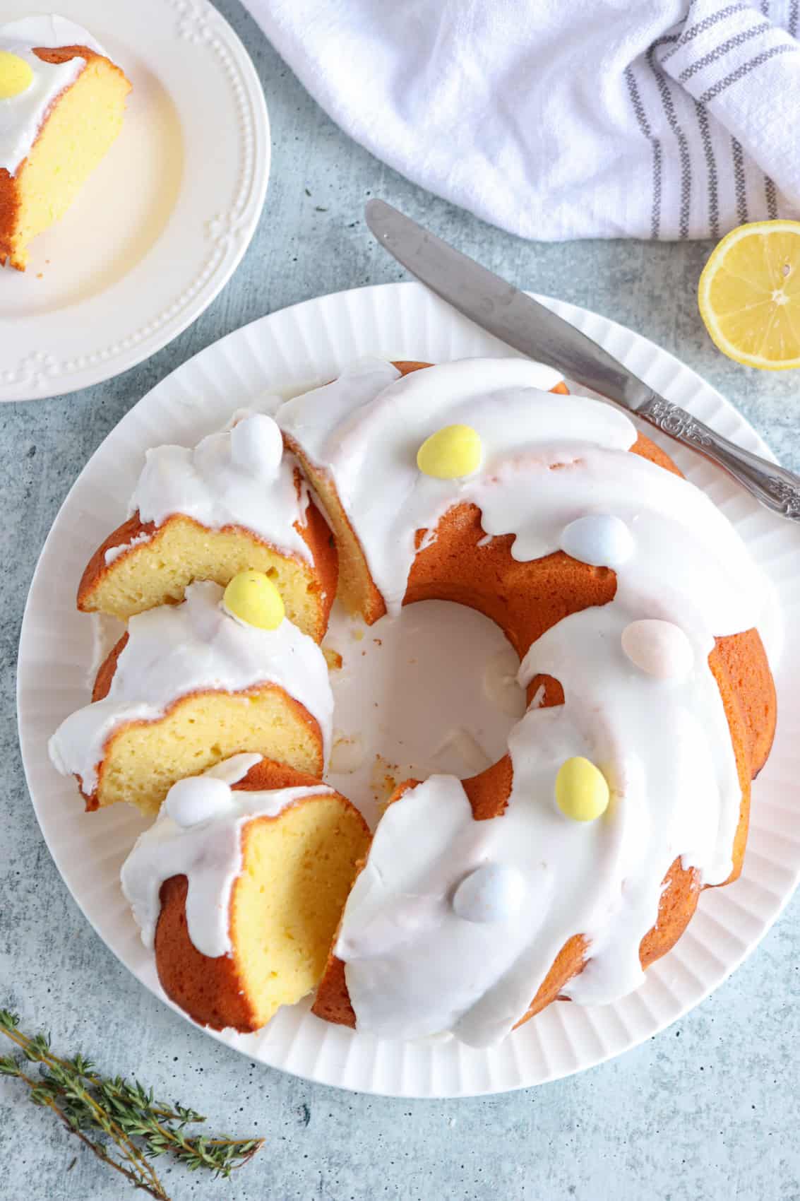 A Polish babka recipe bundt cake with white icing and pastel candy eggs on top, displayed on a white plate. A knife, lemon slice, and a plate with a cake slice are nearby, with a striped towel in the background.