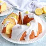 A lemon babka cake with white icing, garnished with pastel candy eggs, sits on a white plate. A slice is cut and placed on a small plate nearby, while two lemon halves and a striped towel add color.