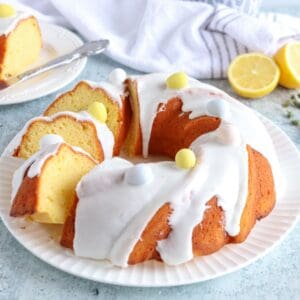 A lemon babka cake with white icing, garnished with pastel candy eggs, sits on a white plate. A slice is cut and placed on a small plate nearby, while two lemon halves and a striped towel add color.