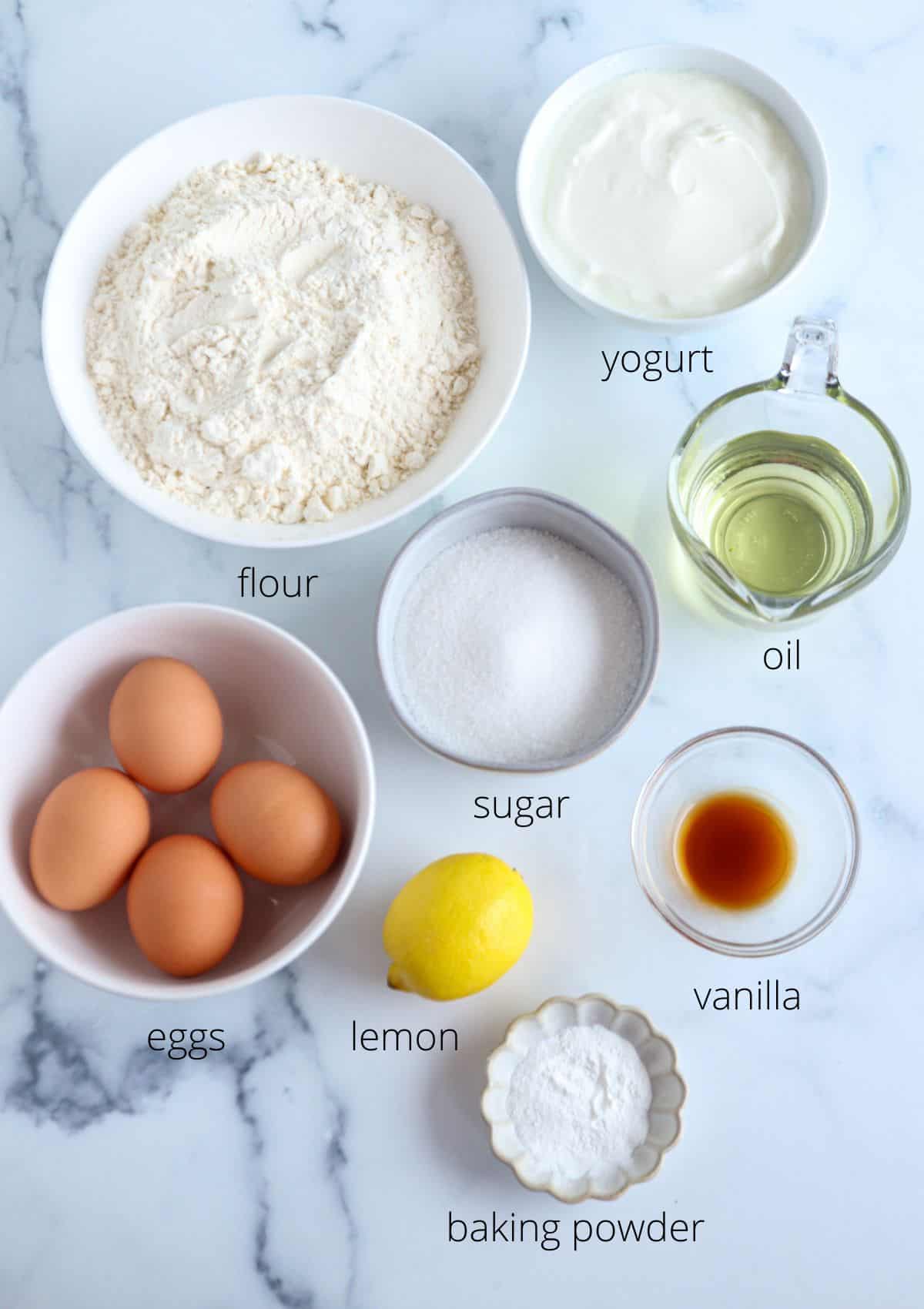 A top-down view of baking ingredients labeled for a Polish babka recipe: a bowl of flour, yogurt, oil, eggs, sugar, lemon, baking powder, and vanilla arranged on a marble surface.