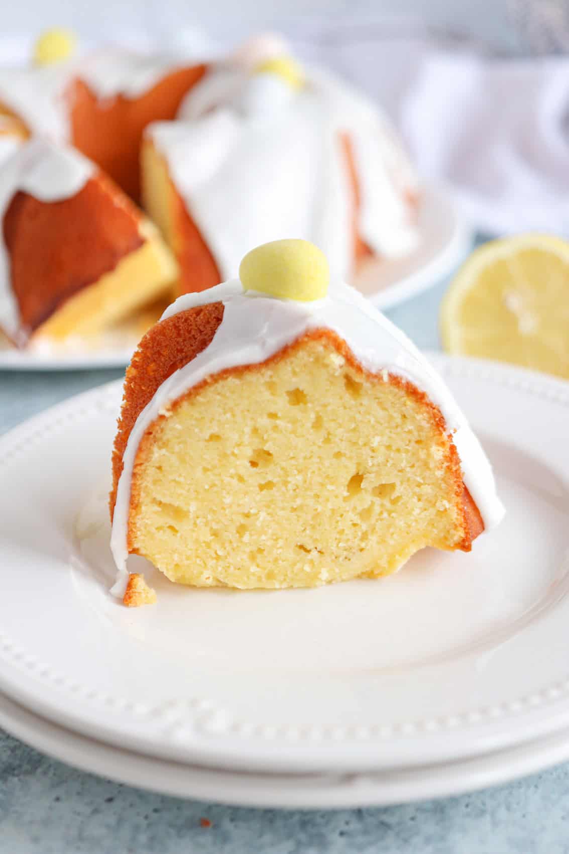 A slice of yellow bundt cake with white icing and a small yellow candy on top, served on a white plate. Inspired by a classic Polish babka recipe, the rest of the cake and a lemon slice are in the background.