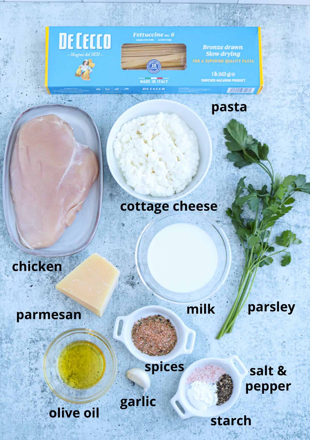 Top view of raw ingredients for high protein chicken Alfredo: boxed fettuccine, raw chicken breast, cottage cheese, parsley, parmesan wedge, olive oil, minced garlic, milk, mixed spices, salt and pepper, and starch.