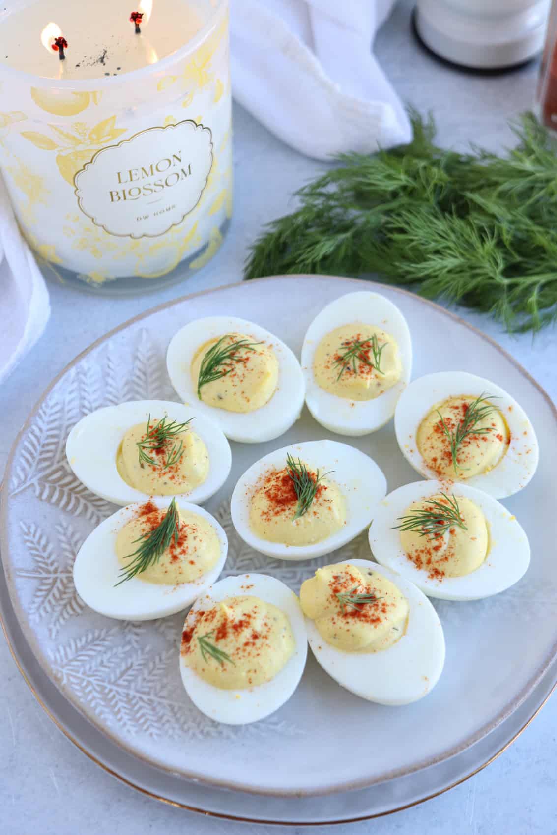 A plate of deviled eggs garnished with fresh dill and paprika sits beside a lit lemon blossom candle, a bunch of dill, and a bowl of ricotta pesto pasta on a light surface.