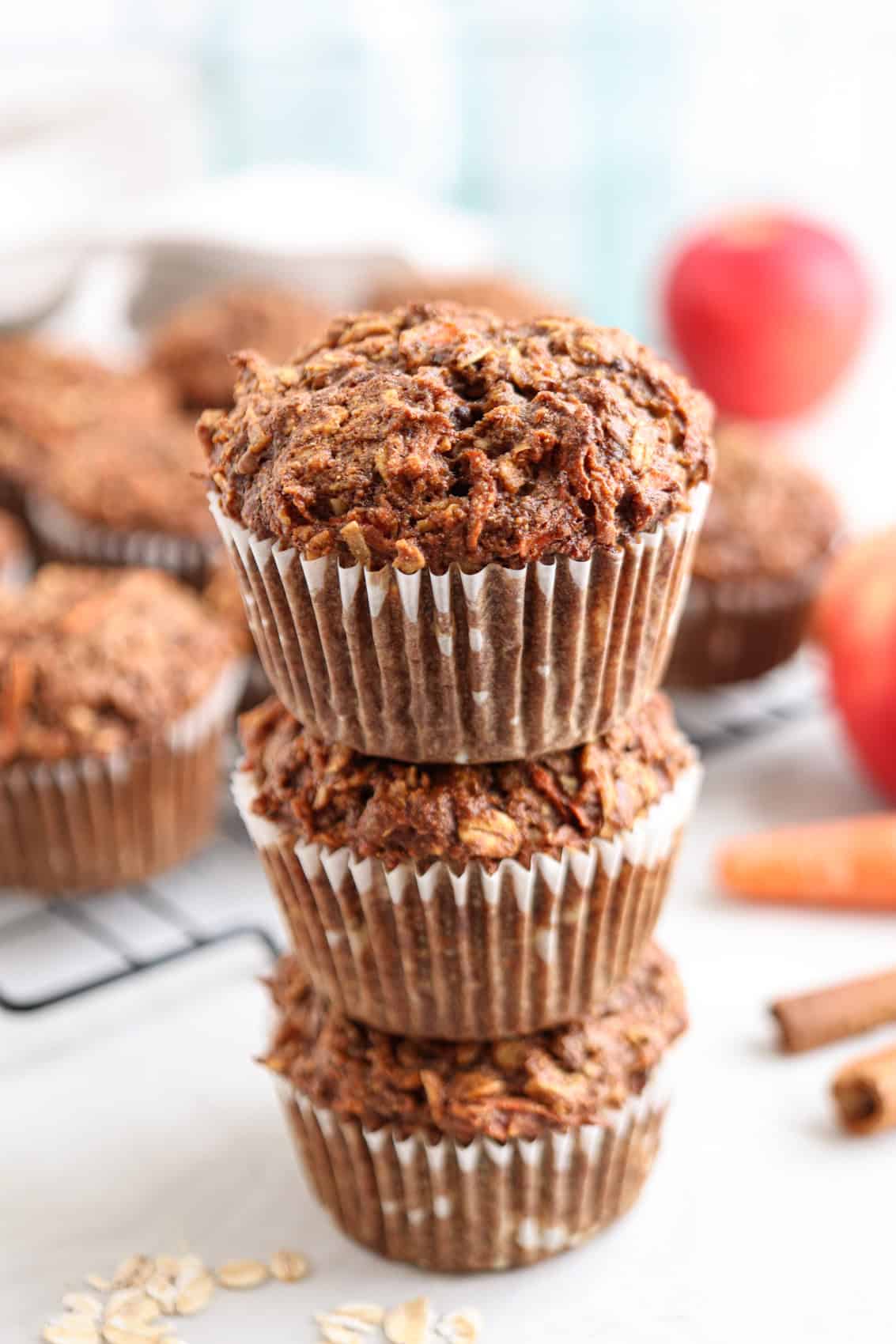 Three healthy morning glory muffins stacked on top of each other, wrapped in paper liners, with more muffins on a cooling rack in the background. Fresh apples, a carrot, oats, and cinnamon sticks are also visible nearby.
