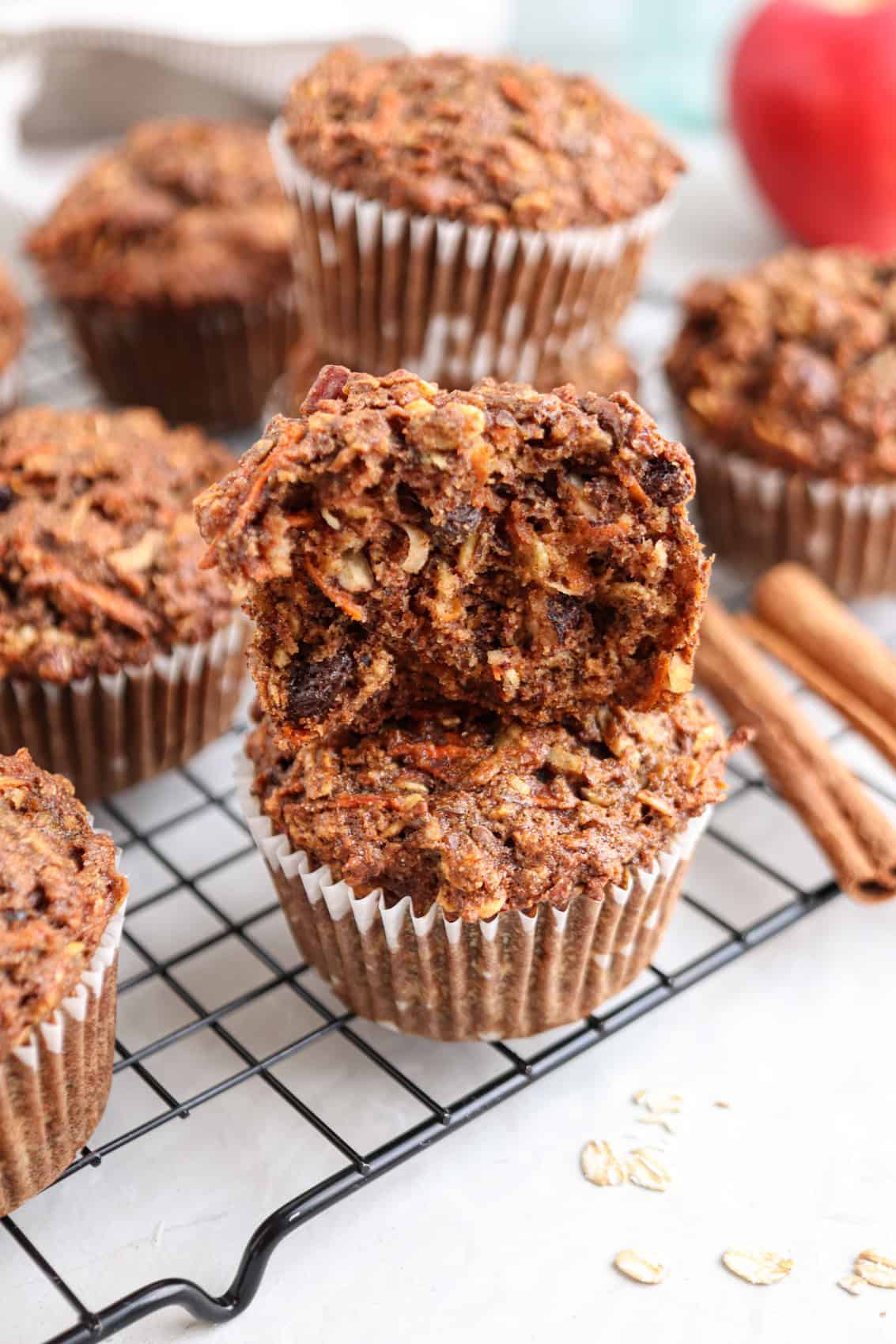 A stack of moist, homemade muffins with a crumbly texture sits on a cooling rack. One muffin is split in half, revealing oats, nuts, and chocolate chips. Two cinnamon sticks and an apple are in the background.