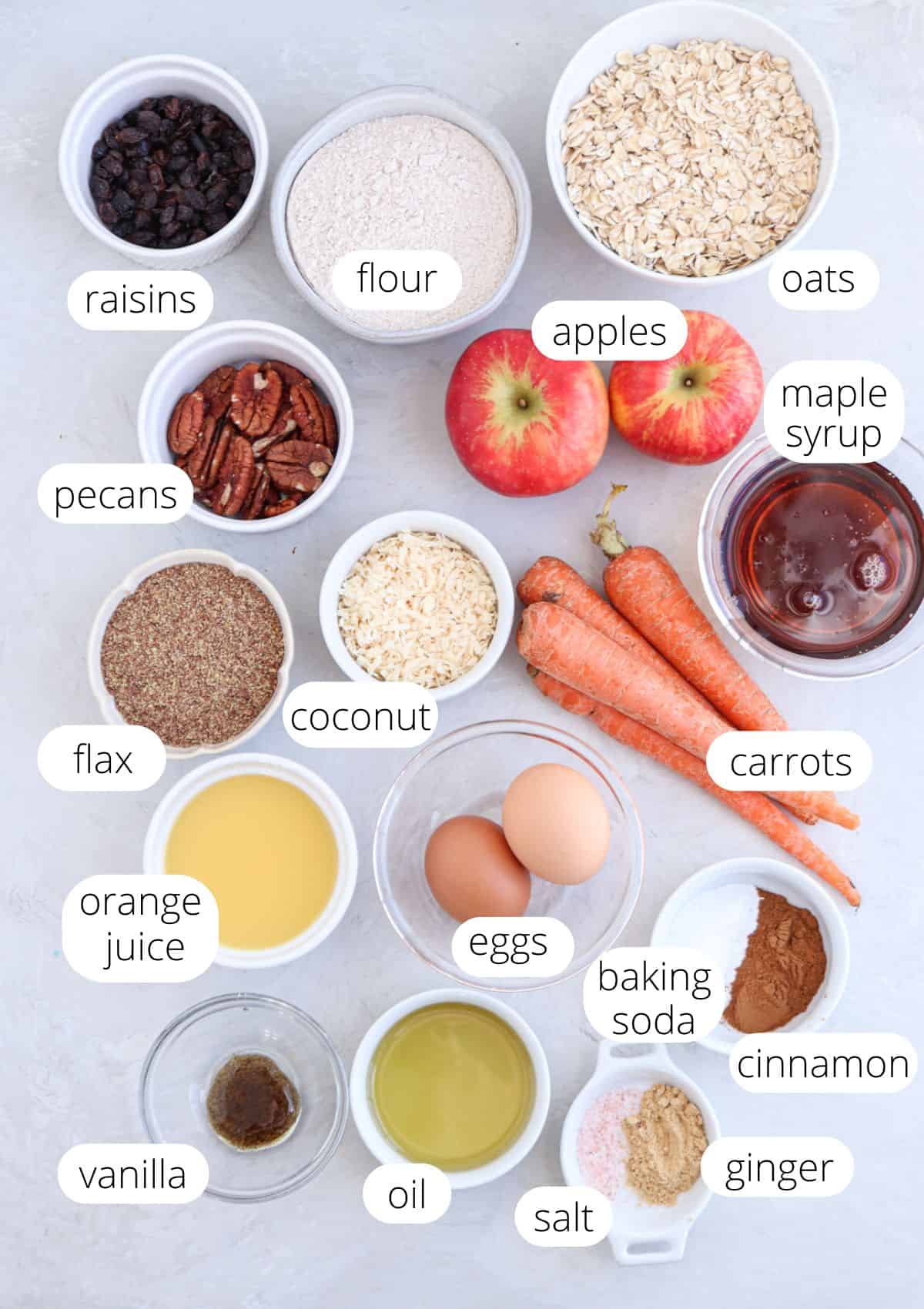 Various labeled bowls and ingredients are arranged on a light surface, including raisins, flour, oats, apples, maple syrup, pecans, flax, coconut, carrots alongside cinnamon and ginger.