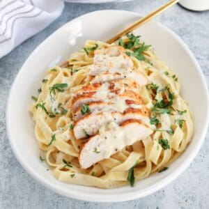 A white bowl filled with high protein chicken Alfredo fettuccine in creamy sauce, topped with sliced grilled chicken breast and garnished with chopped parsley. A gold fork rests on the side of the bowl.