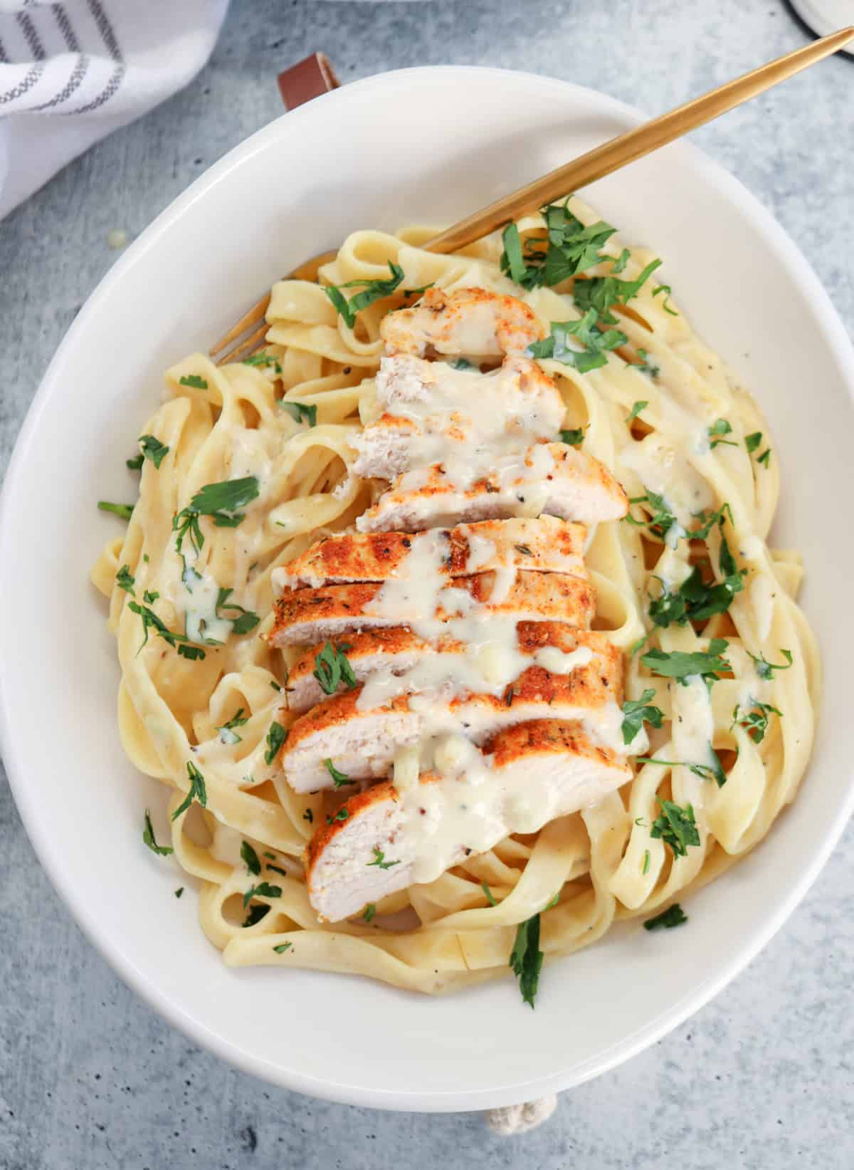 A white bowl of high protein chicken Alfredo fettuccine in creamy Alfredo sauce, topped with sliced grilled chicken breast and garnished with chopped parsley. A gold fork rests on the side of the bowl.