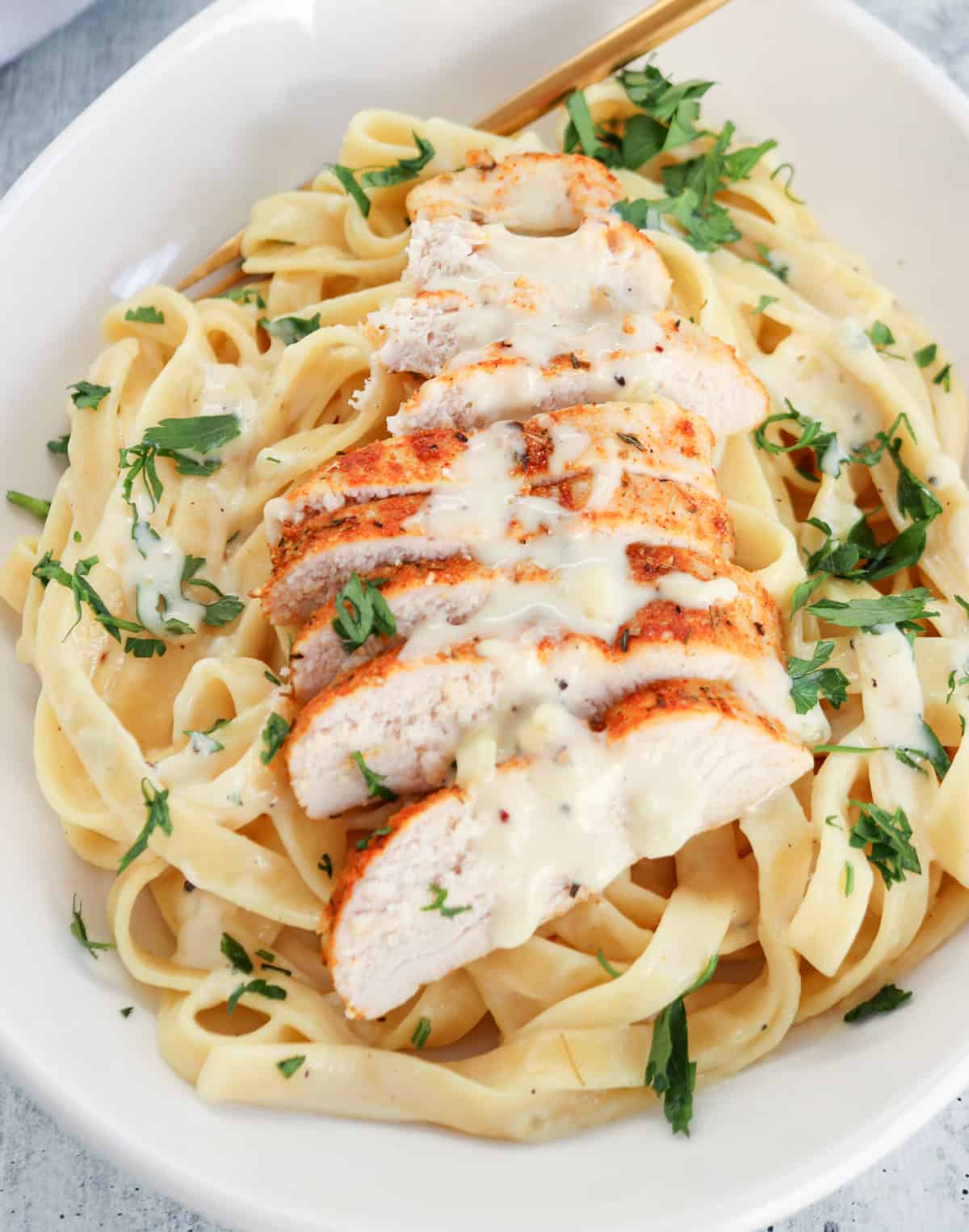 A white bowl filled with high protein chicken Alfredo fettuccine pasta, topped with sliced grilled chicken breast, creamy Alfredo sauce, and garnished with chopped fresh parsley. A gold fork rests on the edge of the bowl.