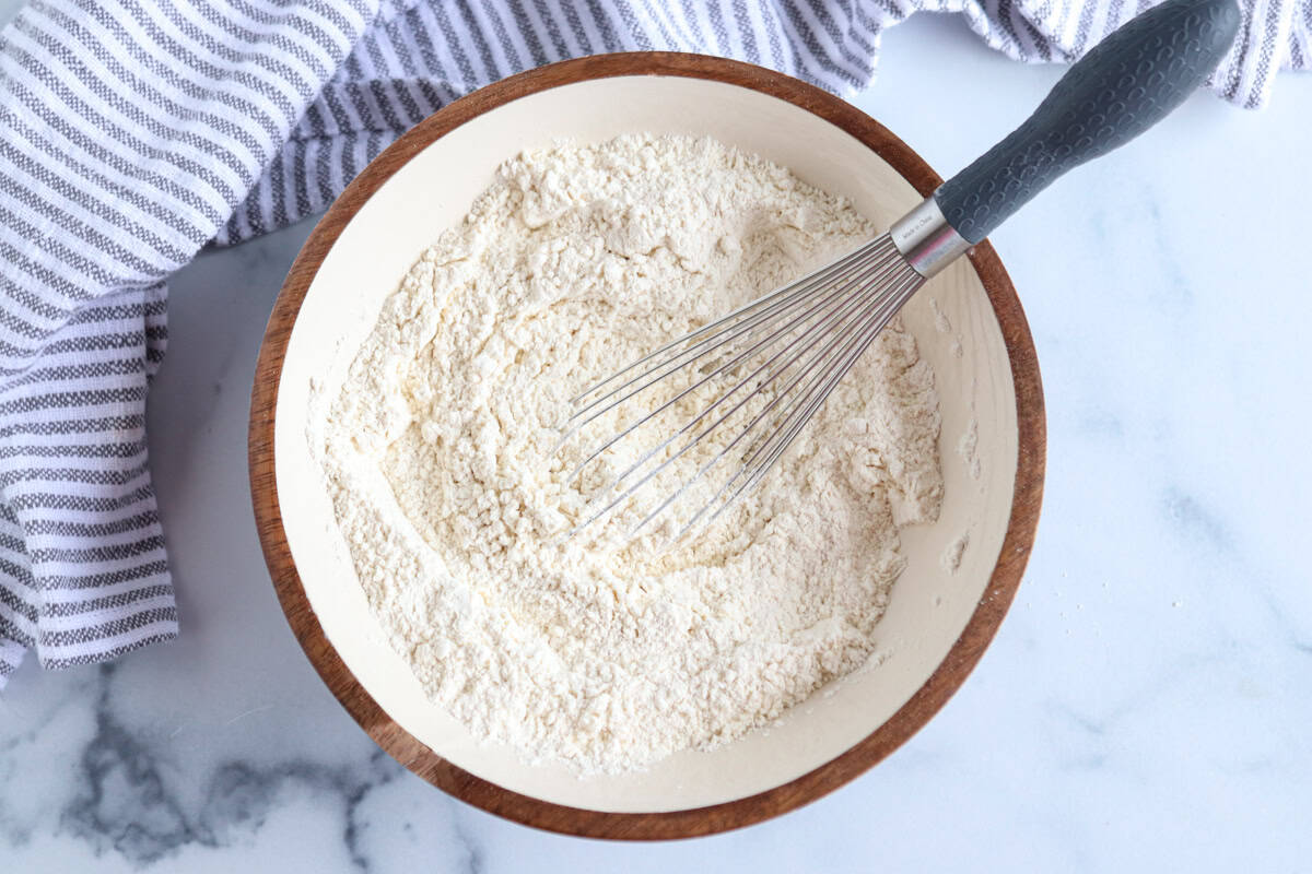 A whisk rests in a wooden bowl filled with flour mixture on a marble surface. A gray and white striped kitchen towel lies nearby.