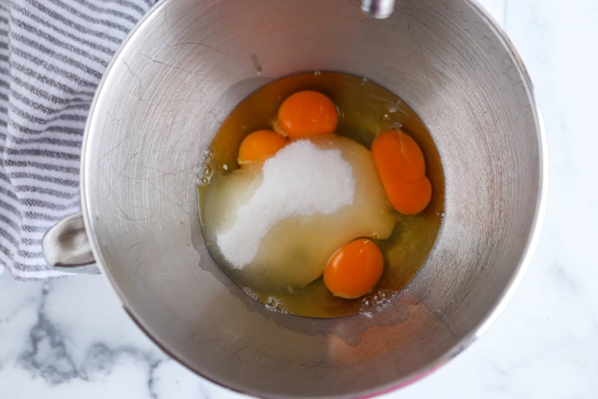 A metal mixing bowl containing four cracked eggs and a mound of sugar sits on a marble countertop with a gray and white striped cloth nearby.