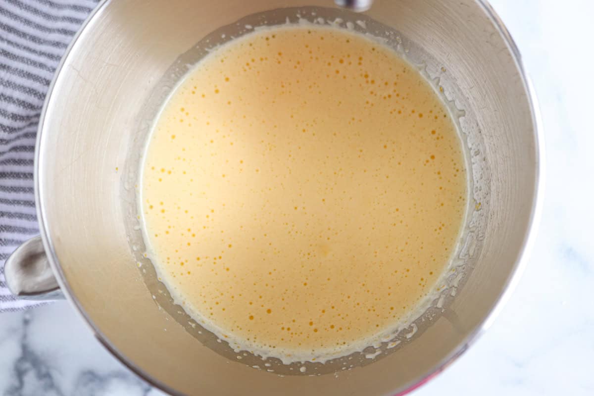 A metal mixing bowl filled with a light yellow, frothy batter sits on a white surface next to a gray and white striped cloth.