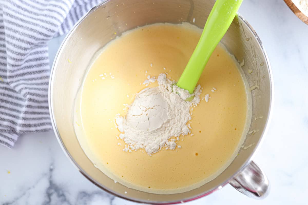 A mixing bowl containing a pale yellow batter with a mound of flour on top, being stirred with a green spatula. A striped towel is next to the bowl on a light marble surface.