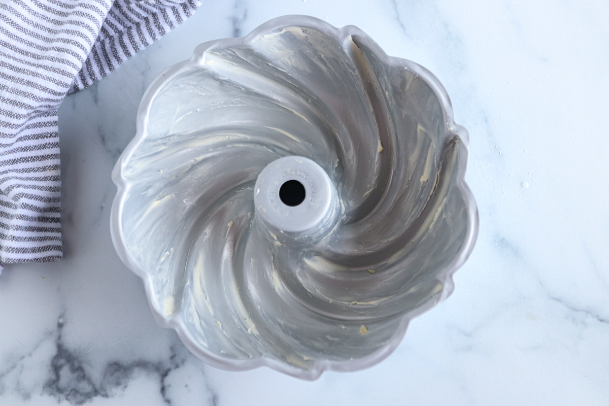 A greased Bundt cake pan with a swirl pattern sits on a white marble surface next to a gray and white striped towel.