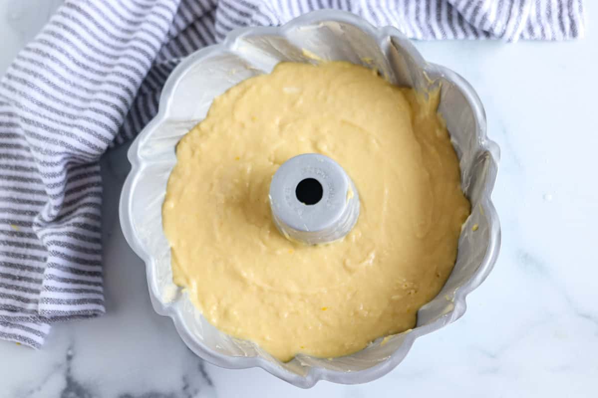 A bundt pan filled with yellow cake batter sits on a white marble surface next to a gray and white striped kitchen towel.