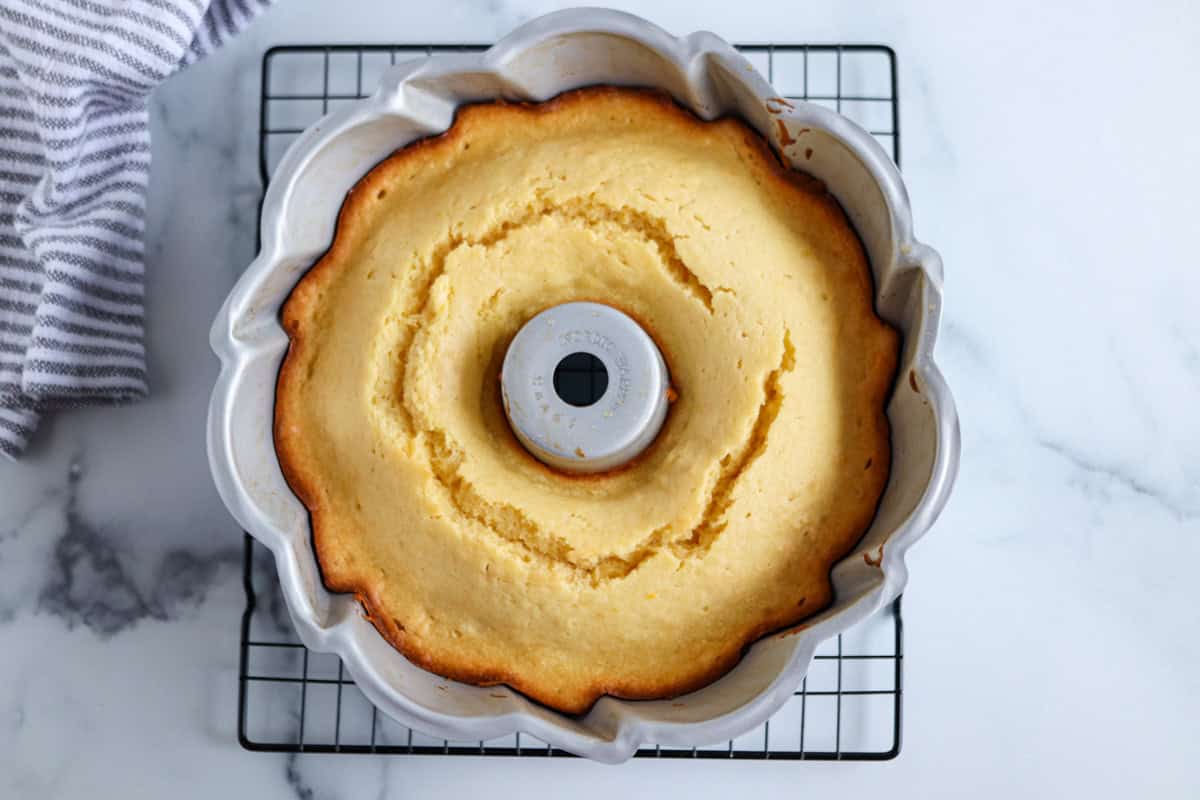 A golden-brown Bundt cake sits in a fluted metal pan on a cooling rack. The cracked ring around the center complements the white marble surface and striped cloth.