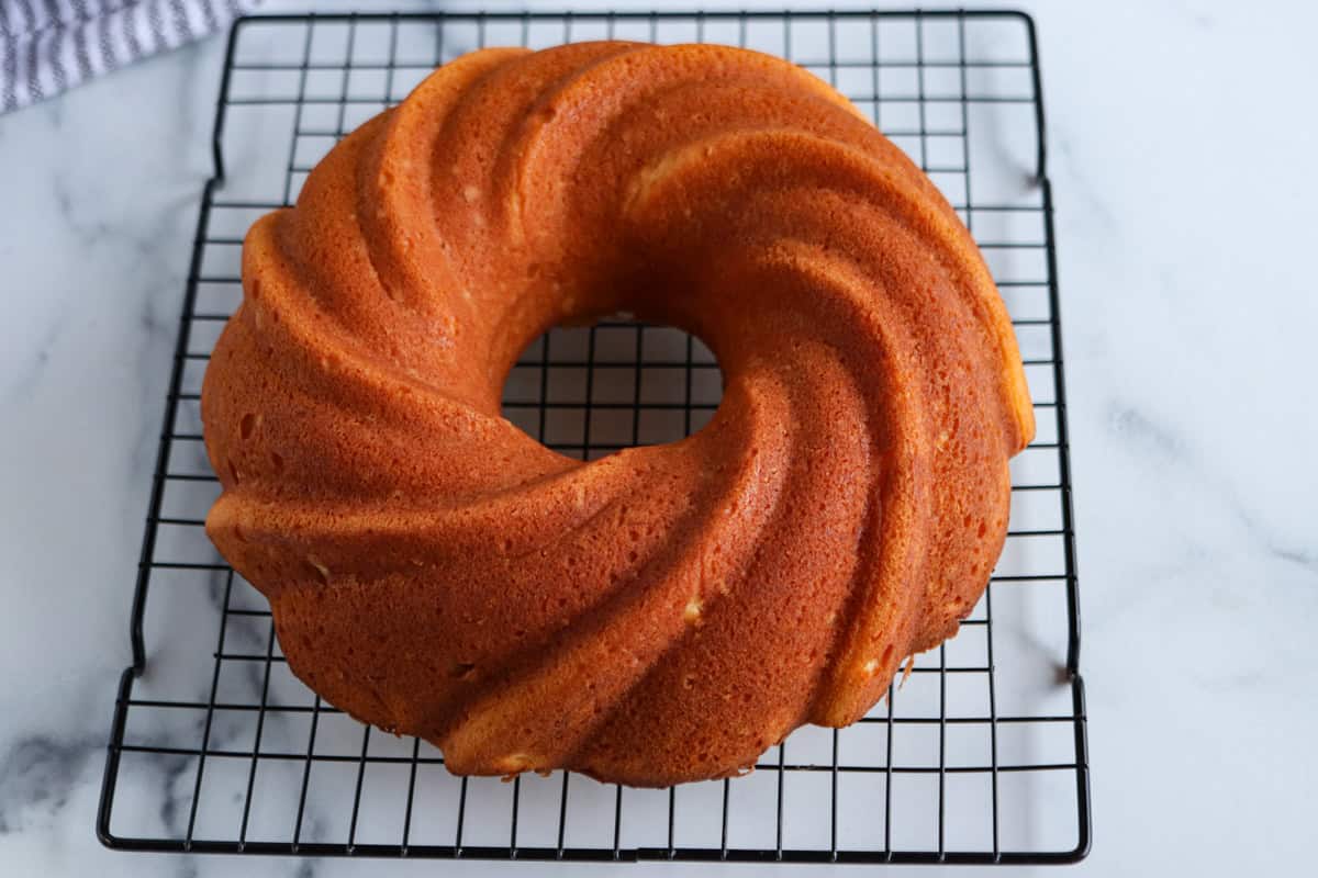 A golden-brown Bundt cake with a swirled pattern sits on a black wire cooling rack.