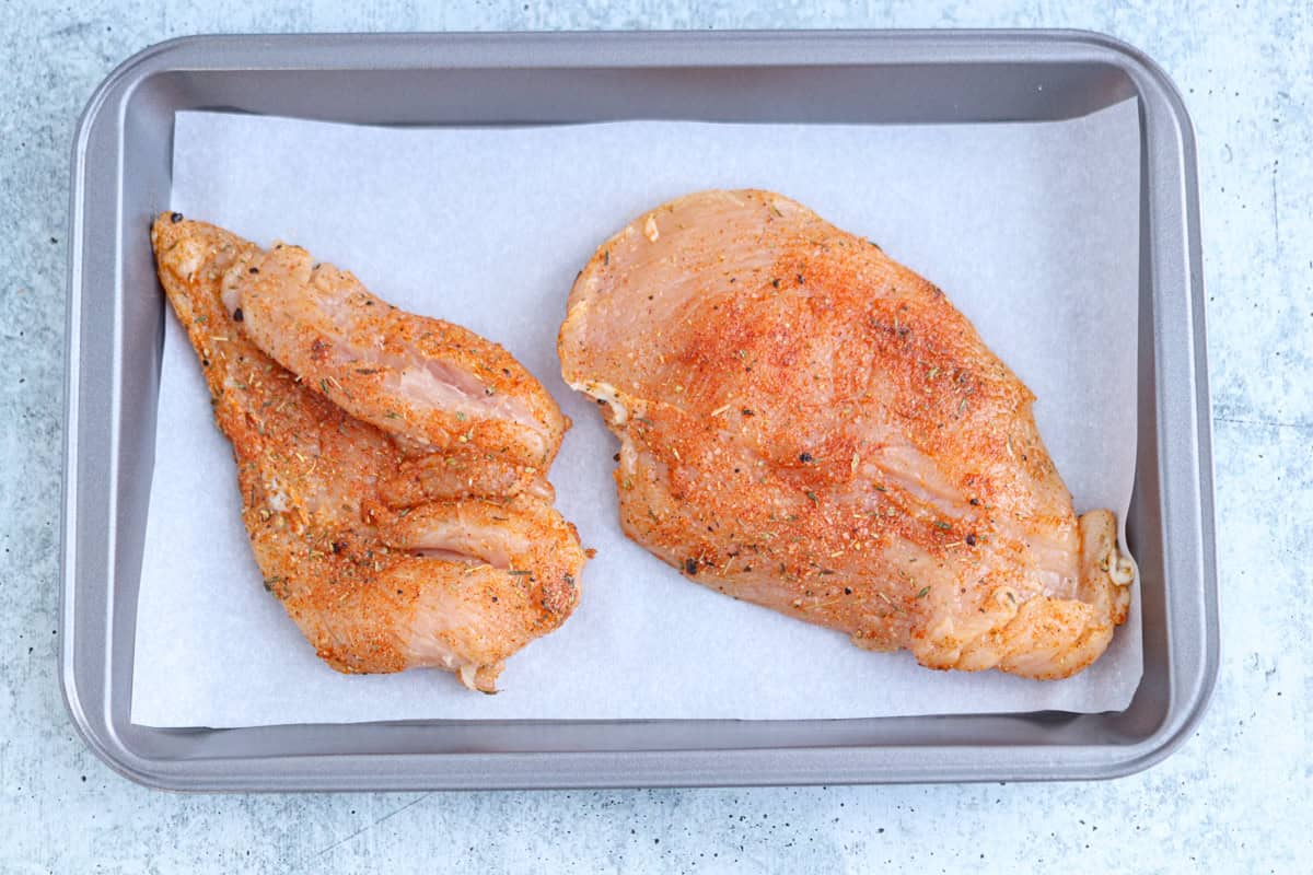 Two seasoned, raw chicken breasts placed on a sheet of parchment paper in a rectangular baking tray, ready for cooking the perfect high protein chicken alfredo.