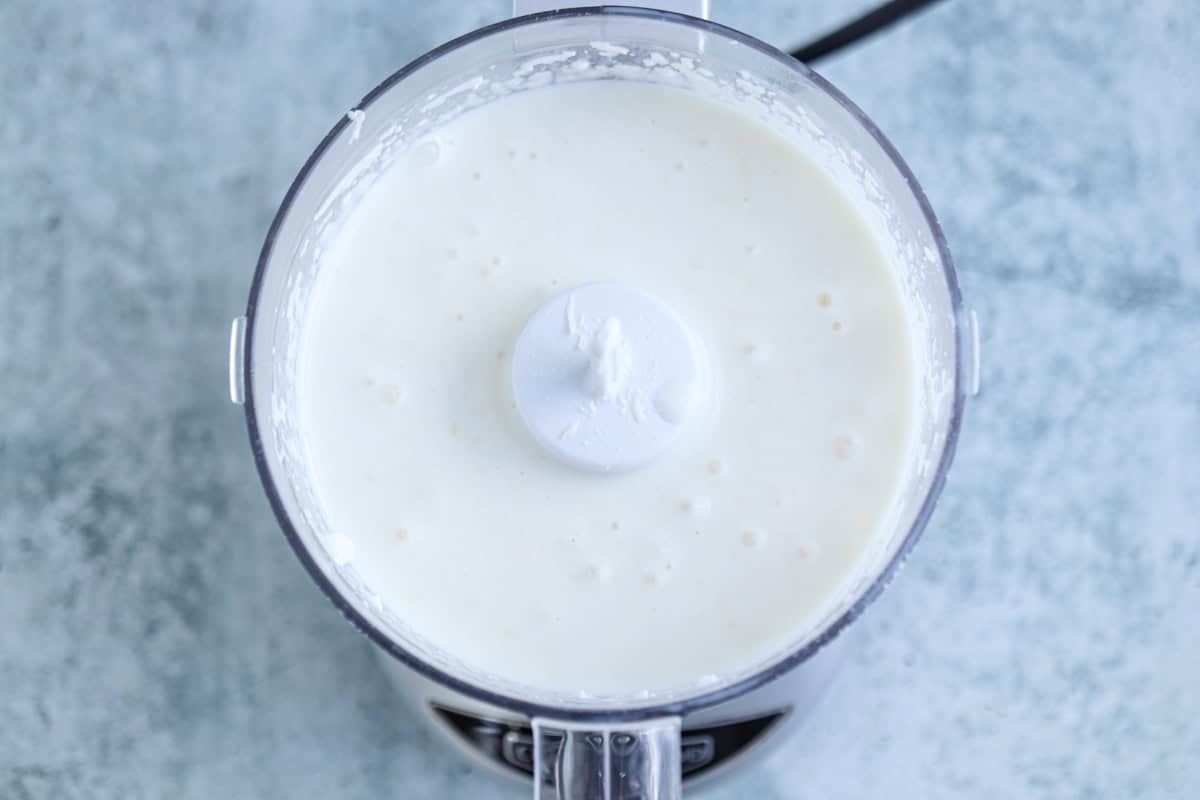 A food processor filled with a smooth, creamy white high protein chicken alfredo mixture, viewed from above on a light gray countertop.