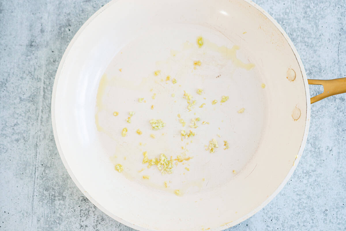 A white frying pan with sautéed minced garlic and oil on the bottom, perfect for starting a high protein chicken alfredo, sits on a light gray countertop.