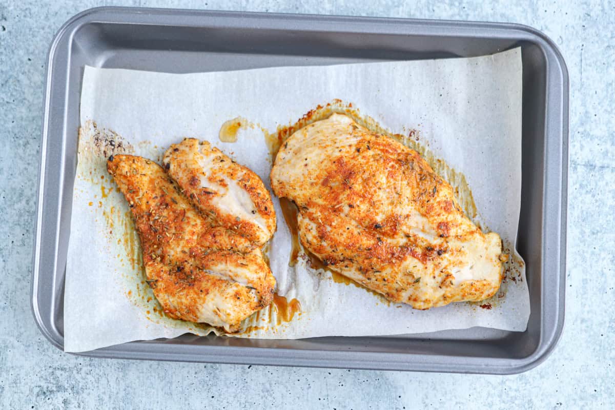 Two cooked, seasoned chicken breasts on parchment paper in a metal baking tray with visible juices—perfect to slice for a high protein chicken alfredo—set against a light gray background.