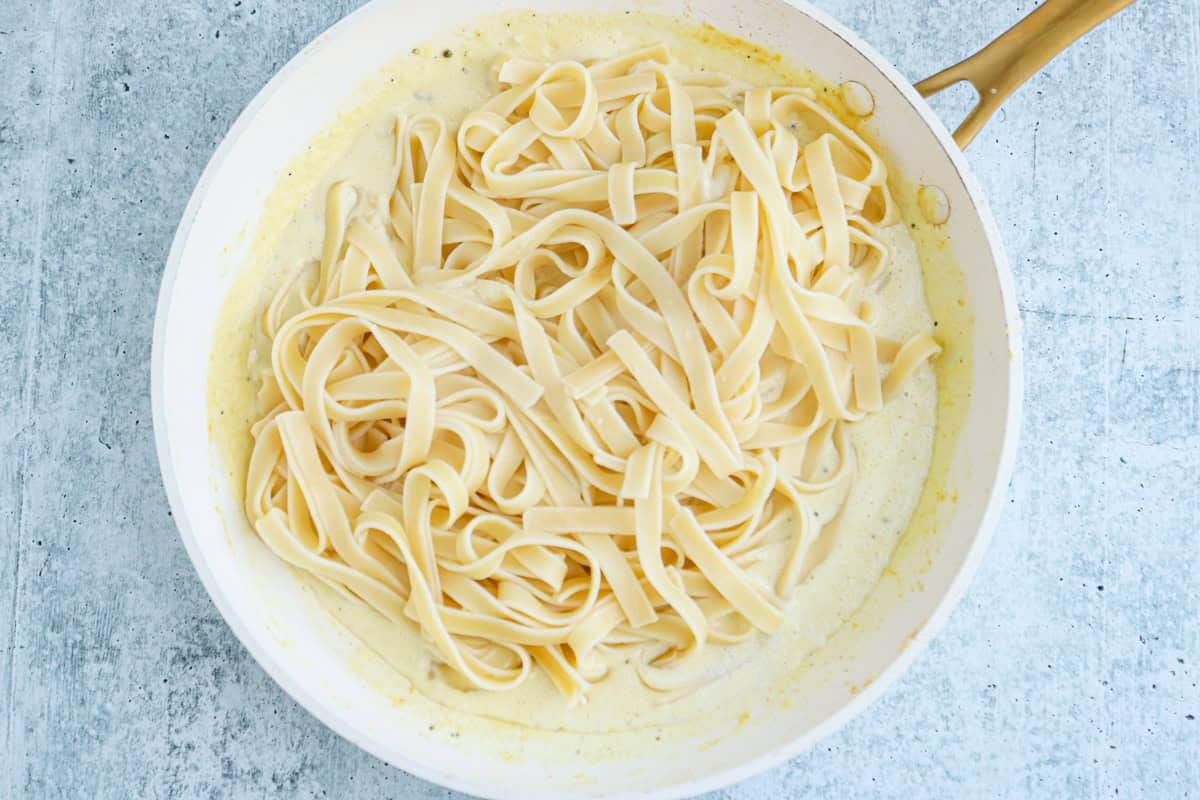 A white skillet with cooked fettuccine pasta in a creamy high protein chicken alfredo sauce, resting on a light gray textured surface.